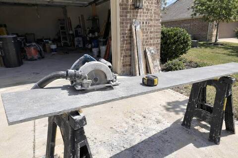 A long gray fiber cement plank on two sawhorses in a driveway, a dusty circular saw with a shroud resting on it and a tape measure nearby