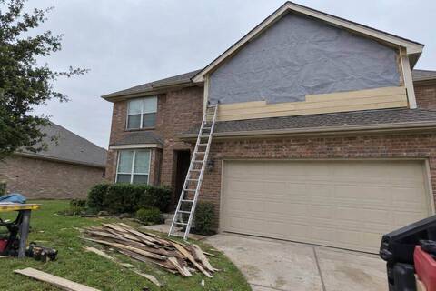 The upper gable of a brick house with old boards removed, gray house wrap exposed and a few new tan planks started along the bottom, seen from the driveway