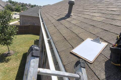 The top of an extension ladder resting against a gutter beside a clipboard on the roof edge, with the shingle field and a vent stack stretching up behind
