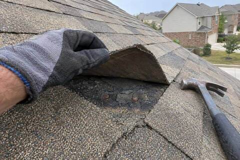 A gloved hand lifting a faded shingle tab to show two rusted nail heads and cracked sealant underneath, on a roof slope in flat daylight