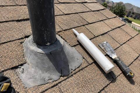 A cracked, sun-bleached rubber collar around a plumbing vent pipe on a shingle roof, a tube of sealant and a putty knife beside it