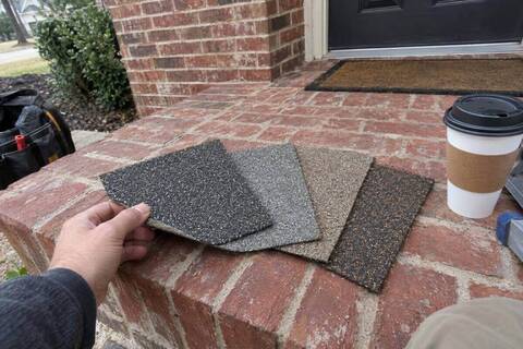 Four shingle samples in different grays and browns fanned out on a brick porch step, one bent back at the corner between a thumb and finger