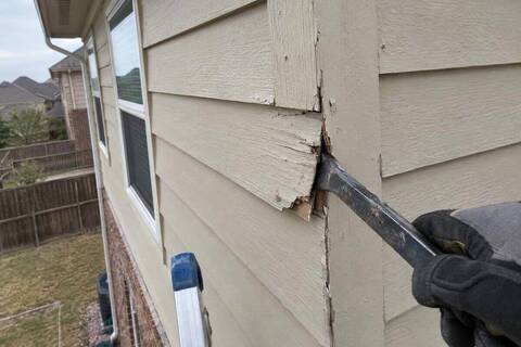 A close view of a split and swollen painted board at an outside corner of upper-story siding, with a flat pry bar hooked behind it
