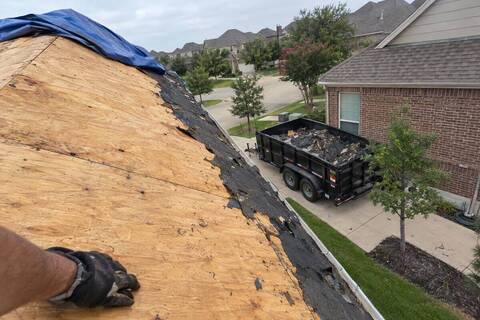 A roof slope stripped to tan wood decking with torn black felt still clinging at the edge, a blue tarp folded over the ridge and a dump trailer parked below in a driveway