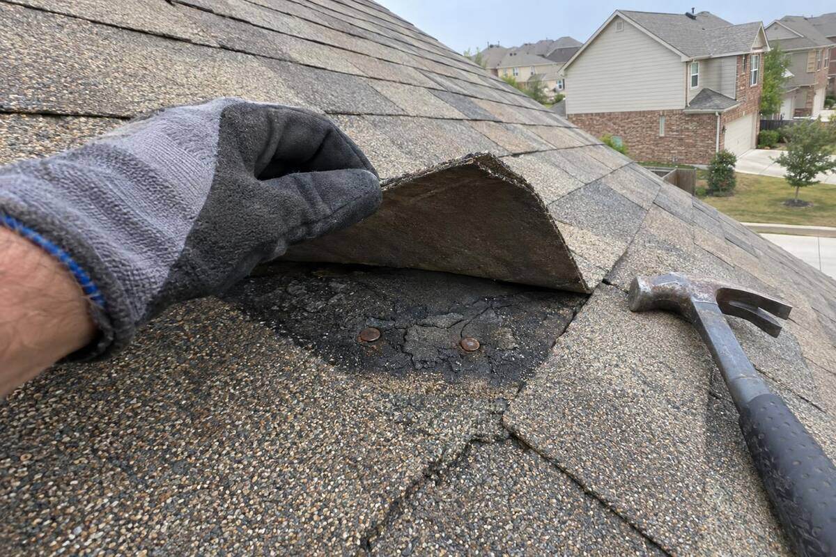 A gloved hand lifting a faded shingle tab to show two rusted nail heads and cracked sealant underneath, on a roof slope in flat daylight