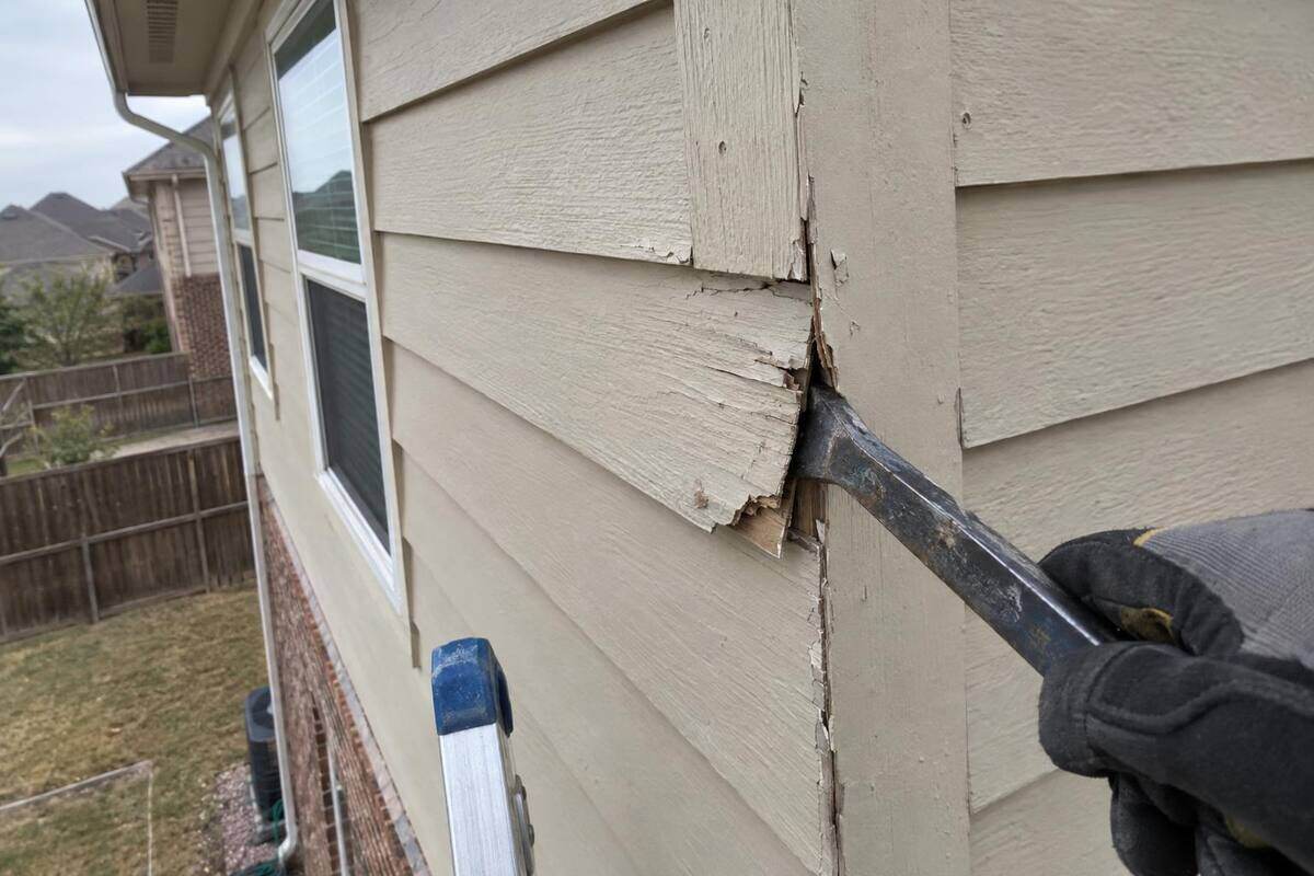 A close view of a split and swollen painted board at an outside corner of upper-story siding, with a flat pry bar hooked behind it